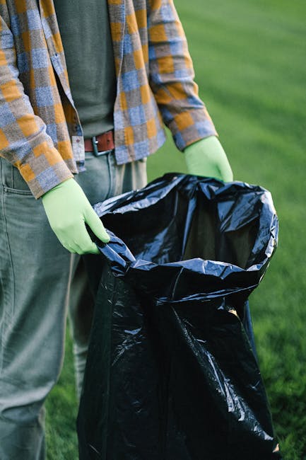 A person standing outdoors on a grassy area, wearing a plaid shirt in shades of blue, yellow, and gray, with green gloves and light-colored trousers secured with a brown belt. They are holding open a large, partially gathered black garbage bag, which appears to be made of glossy plastic material. The background features a lush green lawn which suggests a garden or park setting, and the lighting is natural and evenly diffused. The scene indicates an activity related to waste collection or rubbish disposal, akin to private rubbish removal services offered by companies like Rubbish Removal Highbury, where individuals handle their own waste for local or alternative disposal solutions. The person’s stance and the focus on the trash bag highlight preparation for rubbish collection or clearance, emphasizing the practical aspects of waste management in an outdoor environment, with no visible vehicle or equipment beyond the trash bag and gloves.