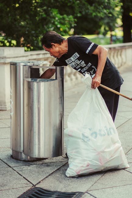 A middle-aged man with dark hair, dressed in a black short-sleeved T-shirt and dark trousers, is seen collecting rubbish into a large white plastic bag. He is using a wooden stick or stick-like tool to help load the bag. The man is standing on a paved urban sidewalk, which appears to be made of concrete slabs, next to a cylindrical public bin made of shiny stainless steel. The bin has a slit on top for easy rubbish disposal and appears to be clean and well-maintained. In the background, there is a stone balustrade or low wall, with lush green trees and out-of-focus shrubbery providing a natural backdrop, suggesting an outdoor setting in a park or city area. The lighting is natural and bright, indicating daytime, with sunlight casting soft shadows. This scene exemplifies a responsible approach to rubbish collection and disposal, aligning with private waste management or rubbish removal services, such as those provided by Rubbish Removal Highbury, highlighting effective on-site clearance of waste without reliance on local authority bins.