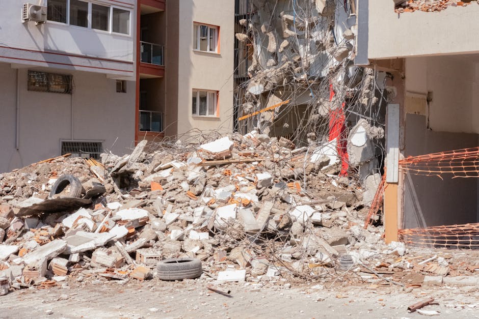 A large pile of construction debris and rubble is situated in front of a multi-storey residential building, with visible windows and balconies. The debris includes broken concrete blocks, bricks, twisted metal rebar, and a scattering of loose wires. Several used car tyres, one black and slightly worn, are partially embedded within the rubble, resting on the uneven surface. To the right, an orange plastic safety barrier is seen, enclosing part of the area, indicating an active or recent demolition or on-site clearance. The building's exterior wall shows damage and missing sections, exposing the interior structure. Natural daylight illuminates the scene, highlighting the rough textures and grey, beige, and reddish tones of the rubble and building materials. This scene depicts a typical setting where a private waste removal service, such as Rubbish Removal Highbury, facilitates the safe clearing of construction waste from urban properties, supporting alternative disposal options outside of municipal collection.