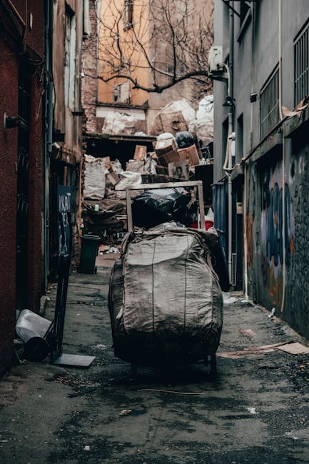 A narrow urban alleyway lined with graffiti-covered walls and cluttered with scattered debris, gravel, and patches of dirt on the ground. In the foreground, a large, grey, weathered garbage bag is attached to a metal trolley or cart, positioned centrally with its top partially open revealing black plastic waste inside. Behind the trolley, an extensive pile of miscellaneous rubbish, including sack bags, cardboard boxes, and broken items, is stacked against a backdrop of bare tree branches and a brick building in the distance. The alley appears somewhat neglected, with signs of staining and dirt on the pavement, and the scene is illuminated by soft natural light, suggesting an overcast day. This image relates to independent waste collection or rubbish removal services, emphasizing the need for professional rubbish clearance in residential or urban environments, as offered by Rubbish Removal Highbury, to manage such clutter effectively.