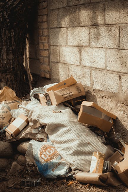 A large pile of construction debris and rubble is situated in front of a multi-storey residential building, with visible windows and balconies. The debris includes broken concrete blocks, bricks, twisted metal rebar, and a scattering of loose wires. Several used car tyres, one black and slightly worn, are partially embedded within the rubble, resting on the uneven surface. To the right, an orange plastic safety barrier is seen, enclosing part of the area, indicating an active or recent demolition or on-site clearance. The building's exterior wall shows damage and missing sections, exposing the interior structure. Natural daylight illuminates the scene, highlighting the rough textures and grey, beige, and reddish tones of the rubble and building materials. This scene depicts a typical setting where a private waste removal service, such as Rubbish Removal Highbury, facilitates the safe clearing of construction waste from urban properties, supporting alternative disposal options outside of municipal collection.