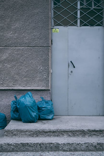 A group of four blue plastic rubbish bags, tightly tied at the top, are stacked on a concrete pavement in front of a grey, textured exterior wall. The bags appear to contain waste and are placed near a light grey metal door with a black door handle and a small lock, positioned on the right side of the image. To the upper right of the door, a section of a window with a diamond-patterned metal grille is visible, suggesting an industrial or commercial building setting. The pavement is slightly raised with three concrete steps leading up to the door area, and the overall scene is illuminated softly, with natural daylight highlighting the urban environment. This scene exemplifies a typical outdoor waste collection or disposal point, possibly managed by an independent rubbish removal service such as Rubbish Removal Highbury, reflecting alternative waste handling methods outside regular municipal collection. The visual focus is on the waste bags and their environment, illustrating a typical scene of private rubbish storage outside a commercial or residential property.
