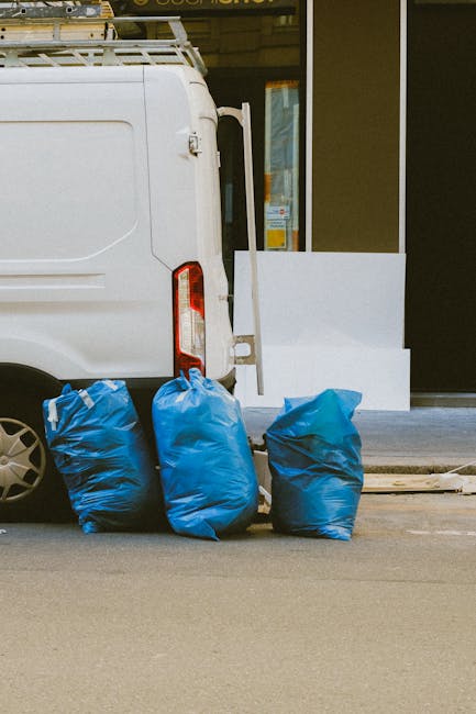 The image shows the rear side of a white commercial van parked on a street, with three blue plastic rubbish bags placed on the ground beside the vehicle. The bags are slightly crumpled and appear full, ready for collection or disposal. The van is positioned close to the curb, with part of a building with large glass windows and a dark door visible in the background. The environment suggests an urban setting, possibly outside a retail or commercial property. The scene reflects a typical scenario of private or independent waste collection, often associated with rubbish removal services like Rubbish Removal Highbury, which handle on-site clearance of rubbish from shops and offices. The lighting is natural, and the overall tone is neutral and professional, focusing on the practical aspect of waste management and rubbish disposal in a non-residential context, emphasizing the visual connection to services that support alternative waste handling outside of local authority rubbish collection.