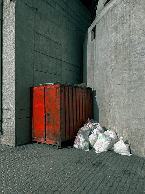 A group of four blue plastic rubbish bags, tightly tied at the top, are stacked on a concrete pavement in front of a grey, textured exterior wall. The bags appear to contain waste and are placed near a light grey metal door with a black door handle and a small lock, positioned on the right side of the image. To the upper right of the door, a section of a window with a diamond-patterned metal grille is visible, suggesting an industrial or commercial building setting. The pavement is slightly raised with three concrete steps leading up to the door area, and the overall scene is illuminated softly, with natural daylight highlighting the urban environment. This scene exemplifies a typical outdoor waste collection or disposal point, possibly managed by an independent rubbish removal service such as Rubbish Removal Highbury, reflecting alternative waste handling methods outside regular municipal collection. The visual focus is on the waste bags and their environment, illustrating a typical scene of private rubbish storage outside a commercial or residential property.