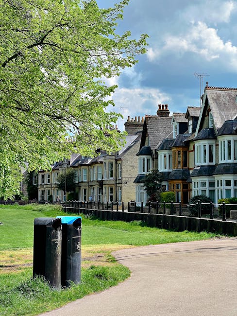 A row of traditional Victorian-style terraced houses with bay windows and chimney stacks, situated along a tree-lined residential street under a partly cloudy sky. In the foreground, two black and blue waste bins are positioned on a paved pathway, with a neatly maintained grassy area and a small wooden fence separating the pavement from the front gardens of the houses. Mature trees with fresh green leaves extend their branches over the grassy verge, providing shade and adding natural greenery to the scene. The environment appears quiet and well-kept, typical of an urban neighbourhood that might employ private waste collection services such as those offered by Rubbish Removal Highbury, especially in the context of adhering to local rules for household rubbish disposal. The overall atmosphere suggests a clean, residential area where external rubbish collection can be managed through private or independent means rather than relying solely on municipal services.