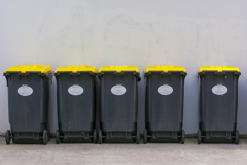 A row of five large wheeled rubbish bins positioned on a concrete pavement against a plain, light-colored wall. The bins are made of dark grey plastic with textured surfaces and feature bright yellow lids that are closed. Each bin has a white oval label with a unique barcode and printed identification numbers, clearly visible on the front. The bins are aligned neatly, with the first and last bins slightly apart from the others. The environment appears to be outdoors, possibly outside a residential or commercial property, with ample natural light illuminating the scene. The setting suggests a typical waste collection area, reflecting a private disposal method that could relate to on-site clearance or alternative rubbish removal services offered by Rubbish Removal Highbury. The overall scene emphasizes organized waste storage, ready for collection or transfer, consistent with professional rubbish management practices common in London boroughs like Islington, where residents might require compliance with local permits or private disposal options.