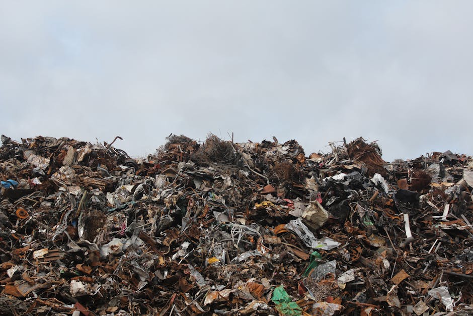 A large mound of mixed waste materials, including crushed and crumpled plastics, metal scraps, cardboard, and other debris, piled high outdoors under a cloudy sky. The rubbish appears densely compacted, with various textures and colors ranging from dark browns and grays to hints of brighter plastics and metallic finishes. The top of the waste heap is irregular, with loose items and debris protruding randomly, indicating recent accumulation or rearrangement. The scene is set on a paved or concrete ground, with the waste heap occupying most of the foreground and extending toward the horizon. The overcast sky provides diffuse lighting, highlighting the rough textures and cluttered appearance of the refuse. This image exemplifies a typical unmanaged rubbish pile, suggesting a need for professional waste collection and disposal services, such as those provided by Rubbish Removal Highbury, which facilitate alternative waste handling and on-site clearance solutions.