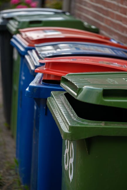 A row of five large wheeled rubbish bins positioned on a concrete pavement against a plain, light-colored wall. The bins are made of dark grey plastic with textured surfaces and feature bright yellow lids that are closed. Each bin has a white oval label with a unique barcode and printed identification numbers, clearly visible on the front. The bins are aligned neatly, with the first and last bins slightly apart from the others. The environment appears to be outdoors, possibly outside a residential or commercial property, with ample natural light illuminating the scene. The setting suggests a typical waste collection area, reflecting a private disposal method that could relate to on-site clearance or alternative rubbish removal services offered by Rubbish Removal Highbury. The overall scene emphasizes organized waste storage, ready for collection or transfer, consistent with professional rubbish management practices common in London boroughs like Islington, where residents might require compliance with local permits or private disposal options.