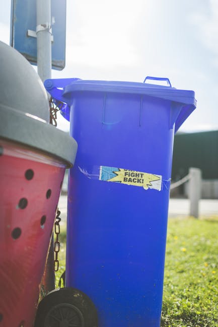 A tall, blue plastic rubbish bin with a slightly textured surface and a closed lid, positioned on a paved or grassy area outdoors. The bin features a small, rectangular sticker with the text 'FIGHT BACK!' in black letters on a yellow background with jagged edges, located near the middle of the front face. The bin is situated next to a red, plastic container with holes and a metal handle, partially visible on the left side of the image. In the background, a metal chain and post barrier can be seen along with a glimpse of a blue signpost on the left, and the scene is illuminated by bright, natural daylight. The setting suggests a public space or roadside environment, aligning with private or alternative waste disposal practices managed by local waste removal services such as Rubbish Removal Highbury.
