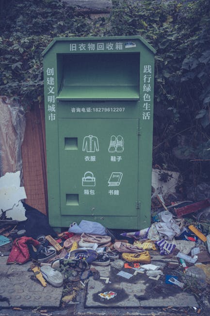 A green recycling and donation collection box with Chinese characters on its surface is positioned outdoors against a backdrop of foliage and wooden fencing. The rectangular metal box features a large central opening at the top, used for depositing clothing, shoes, bags, and books, which are represented by simple icons on the front panel. Surrounding the box, there is a substantial amount of clutter, including discarded shoes, clothing items, and various trash scattered on the ground, indicating neglect or improper disposal. The environment appears as an untidy area possibly meant for waste segregation, yet it is overwhelmed with loose rubbish surrounding the container. The scene highlights issues related to responsible rubbish handling and the importance of proper disposal practices, which Rubbish Removal Highbury often addresses through alternative waste management options for private collection and on-site clearance services in the Highbury area.