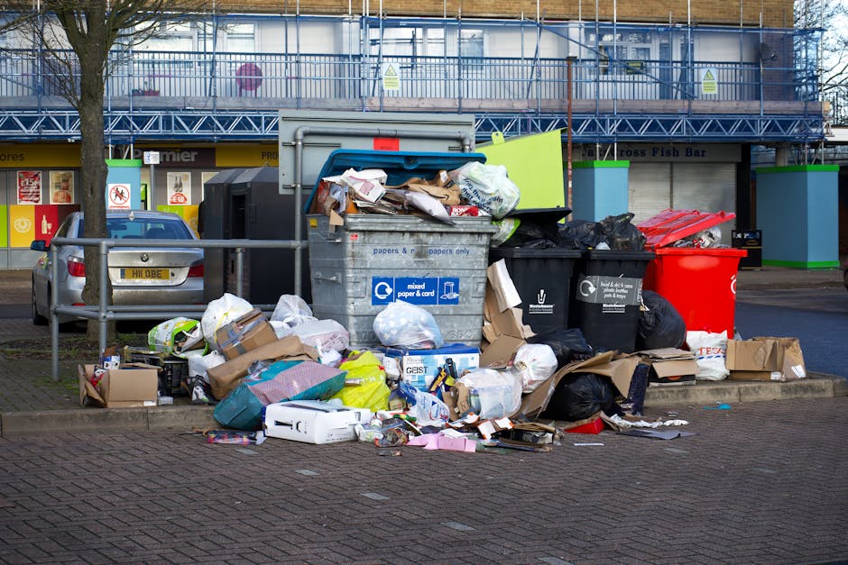 A large pile of mixed household waste including crumpled paper, cardboard boxes, plastic bags, and discarded packaging spread across a paved area next to a metal railing. Several black, red, and grey rubbish bins are situated behind the overflowing trash, with some lids open revealing additional waste. The scene is set in an urban outdoor environment near a parking lot, with a parked car visible on the left side. In the background, there is a building with a blue metal staircase, green-framed shopfronts, and signage indicating retail premises. The waste appears unorganized, with various materials spilling onto the ground, emphasizing an example of illegal dumping or inadequate rubbish collection. The overall atmosphere is neutral and observational, suitable as an illustration of waste disposal challenges that private rubbish removal services like Rubbish Removal Highbury aim to address through alternative waste handling solutions.