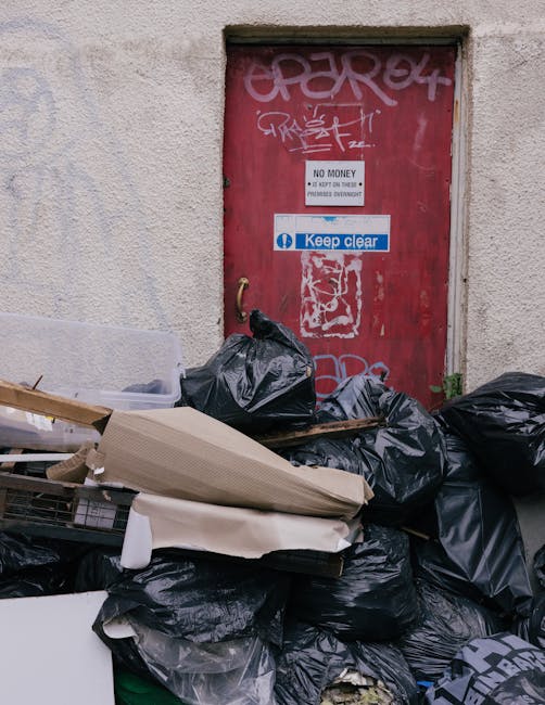 A narrow urban alleyway cluttered with overflowing rubbish bags and scattered debris, including a large, weathered black garbage bag and a metal shopping cart filled with trash. Behind the cart, there is a pile of cardboard boxes, plastic bags, and other waste materials stacked against a wall at the back of the alley. The ground surface appears dirty and stained, with patches of mud and scattered litter. On the left side, a metal fence and a small trash can are visible, while the right side features a building with graffitied walls, an air conditioning unit, and metal security bars on the windows. Overhead, leafless tree branches stretch across the scene, and the overall atmosphere suggests neglect and urban waste accumulation. The natural lighting is diffuse, typical of an overcast day, emphasizing the neglected state of this alleyway where private waste collection might be a consideration. The scene relates to rubbish removal services, illustrating a case of significant waste requiring professional intervention, such as that offered by Rubbish Removal Highbury in the local context of alternative waste handling outside of municipal collection.