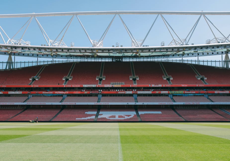 The image shows the interior of a large, modern football stadium with red seating arranged in multiple tiers, some of which have white signage and advertisements. The seats are evenly spaced and form the shape of a recognizable logo or symbol on the central section. The playing field in the foreground has well-maintained green grass with visible mowing patterns and a clearly marked white halfway line. Above the stands, an intricate white steel framework supports a partially covered roof structure, which extends outward over the seating areas. The open sky is visible in the background, indicating daytime with bright, natural lighting illuminating the scene. The stadium appears to be empty, with no spectators or players visible, emphasizing the scale and design of the sports venue, which is suitable for hosting large-scale football events and emergency or private event planning related to grounds maintenance or stadium management by companies such as Rubbish Removal Highbury for waste management or site clearance purposes during events or renovations.