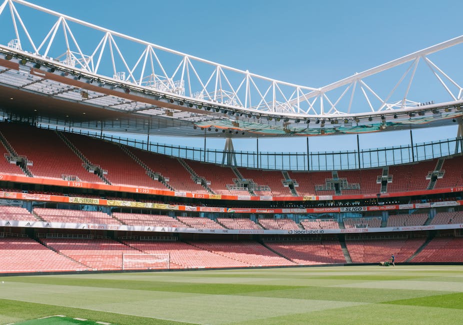 Inside a large, modern stadium with a partially covered roof structure supported by white steel trusses set against a clear, bright blue sky. The interior seating area features multiple tiers of red seats arranged in curved rows, with some sections sectioned off by black barriers. The field below has well-maintained green grass with clearly visible mowing patterns, and a lone individual can be seen working or inspecting the pitch near one corner. The stadium's interior design includes advertising banners along the perimeter of the seating area, with some text visible but not legible. The overall scene emphasizes the expansive, clean, and organized environment typical of a professional sports venue, providing an appropriate context for private waste handling or site clearance activities, as would be performed by a rubbish removal service such as Rubbish Removal Highbury.