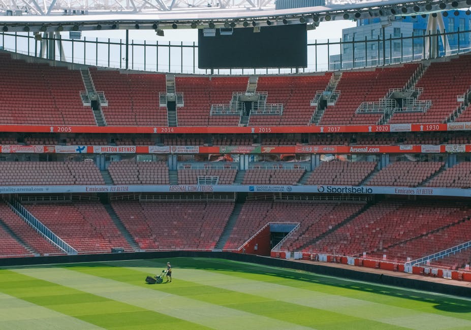 The image shows the interior of a large, modern football stadium with red seating arranged in multiple tiers, some of which have white signage and advertisements. The seats are evenly spaced and form the shape of a recognizable logo or symbol on the central section. The playing field in the foreground has well-maintained green grass with visible mowing patterns and a clearly marked white halfway line. Above the stands, an intricate white steel framework supports a partially covered roof structure, which extends outward over the seating areas. The open sky is visible in the background, indicating daytime with bright, natural lighting illuminating the scene. The stadium appears to be empty, with no spectators or players visible, emphasizing the scale and design of the sports venue, which is suitable for hosting large-scale football events and emergency or private event planning related to grounds maintenance or stadium management by companies such as Rubbish Removal Highbury for waste management or site clearance purposes during events or renovations.